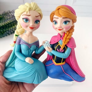 May include: Two figurines of animated characters, one with blonde hair in a braid wearing a blue dress, and the other with auburn hair in a braid wearing a pink cape and blue dress. Both figures are kneeling and holding hands.