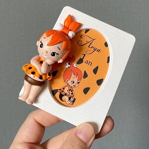 Könnte beinhalten: Ein weißer Rahmen mit einem ovalen Ausschnitt, der ein Cartoon-Bild eines Mädchens mit orangefarbenem Haar und einem braunen und orangefarbenen Tierdruckkleid zeigt. Der Text "Arya 1 an" ist in den ovalen Ausschnitt gedruckt.