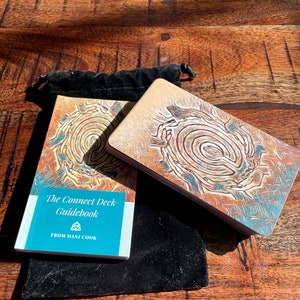 May include: A blue and white guidebook titled "The Connect Deck Guidebook from Dani Cook" with a triquetra symbol. The guidebook is sitting on top of a black velvet bag. A card with a colourful illustration of a spiral pattern is visible in the background.