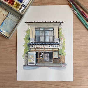 Peut inclure: Aquarelle d'une boutique japonaise avec un distributeur automatique. Le bâtiment a un balcon, un toit bleu foncé et une enseigne avec du texte japonais. Le feuillage vert encadre le bâtiment. Pinceaux et palette d'aquarelle en arrière-plan.