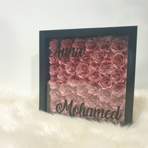 Könnte beinhalten: Eine schwarze, gerahmte Schattenbox, gefüllt mit rosa Papierrosen. Die Namen "Anna" und "Mohamed" sind in schwarzer Schrift auf der Vorderseite der Box geschrieben.