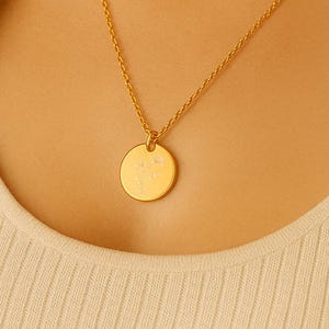 Könnte beinhalten: Eine goldfarbene Halskette mit einem kreisförmigen Anhänger. Der Anhänger hat ein dezentes Design und hängt an einer zarten Kette. Die Halskette wird über einem cremefarbenen Oberteil getragen.