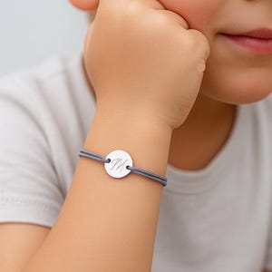 Könnte beinhalten: Ein silbernes Armband mit grauem Band. Das Armband hat einen runden, gravierten Anhänger mit dem Buchstaben "M". Das Armband wird am Handgelenk eines Kindes getragen.