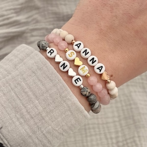 Könnte beinhalten: Drei Armbänder mit weißen Perlen, rosa Perlen und grauen Perlen. Die Perlen sind mit silbernen und goldenen Herzanhängern und Buchstabenperlen durchsetzt, die die Namen "Rinne", "Sime" und "Anna" buchstabieren.