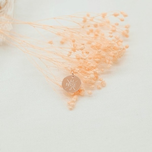 May include: A gold-toned pendant with a compass design and the word "NICE" engraved on it. The pendant is surrounded by dried flowers.
