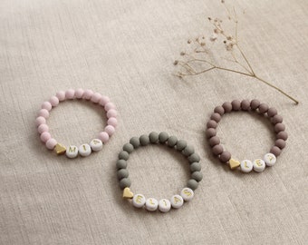 May include: Three personalised beaded bracelets with gold heart charms. The bracelets are pink, grey, and brown. The names on the bracelets are Mia, Elias, and Lea.