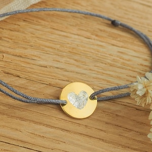 Könnte beinhalten: Ein goldfarbenes Armband mit einem runden Anhänger, der in der Mitte eine silberfarbene, herzförmige Fingerabdruckgravur aufweist. Der Anhänger ist an einer dünnen grauen Kordel befestigt, die durch zwei Löcher an den Seiten verläuft.
