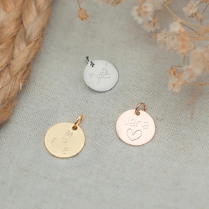 Könnte beinhalten: Drei runde Metallanhänger mit verschiedenen Gravuren. Der Anhänger links ist golden und trägt die Aufschrift "M & G". Der Anhänger in der Mitte ist silberfarben und hat ein handgezeichnetes Design von zwei Händen, die sich halten. Der Anhänger rechts ist roségoldfarben und trägt die Aufschrift "Jana" mit einem kleinen Herz unter dem Namen.