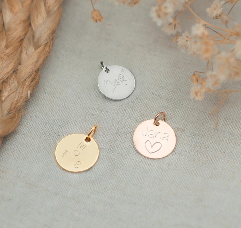 May include: Three round metal charms with different engravings. The charm on the left is gold and says "M & G". The charm in the middle is silver and has a hand-drawn design of two hands holding each other. The charm on the right is rose gold and says "Jana" with a small heart below the name.