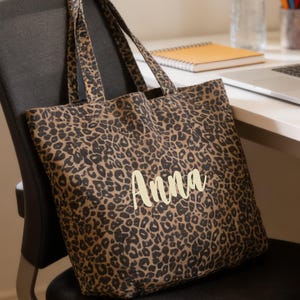 Könnte beinhalten: Eine Tragetasche mit Leopardenmuster und dem Namen "Anna" in hellem Gelb gestickt. Die Tasche liegt auf einem schwarzen Bürostuhl. Ein Laptop, eine Wasserflasche und Buntstifte befinden sich auf dem Schreibtisch im Hintergrund.