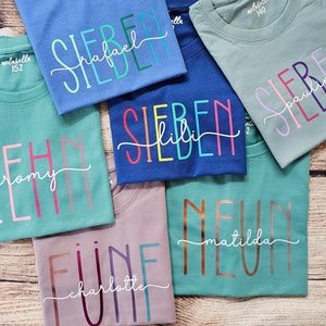 May include: Six short-sleeved cotton t-shirts in various colors including blue, teal, and mauve. Each shirt features a large printed number and a cursive name overlay. The labels inside the collars display the brand name and size numbers.