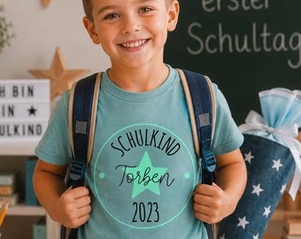May include: A light blue t-shirt featuring a circular graphic with a central green star, the text "SCHULKIND", the name "Torben", and the year "2023". The shirt is worn with a dark blue backpack, and the design is centered on the chest.