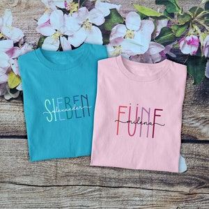 May include: A teal t-shirt with the text "SIEBEN" and "alexander" in cursive, and a light pink t-shirt with the text "F&Uuml;NF" and "milena" in cursive. The teal shirt features multi-colored letters, while the pink shirt has red and purple letters.
