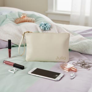 Könnte beinhalten: Cremefarbene Clutch mit dem Aufdruck "Julia", einem goldenen Reißverschluss und einer Handschlaufe. Die Clutch ist von Beauty-Accessoires umgeben, darunter Lipgloss, ein Telefon, Parfüm, eine Haarspange, ein Scrunchie und eine Halskette.