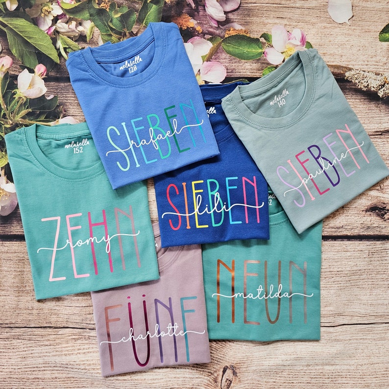 May include: Six short-sleeved cotton t-shirts in various colors including blue, teal, and mauve. Each shirt features a large printed number and a cursive name overlay. The labels inside the collars display the brand name and size numbers.