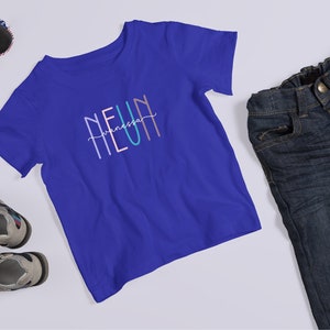 May include: A royal blue short-sleeve t-shirt featuring the text NEUN with the name Vanessa written in cursive script across the center. The shirt is displayed alongside a pair of dark wash denim jeans, gray and black sandals, and sunglasses.