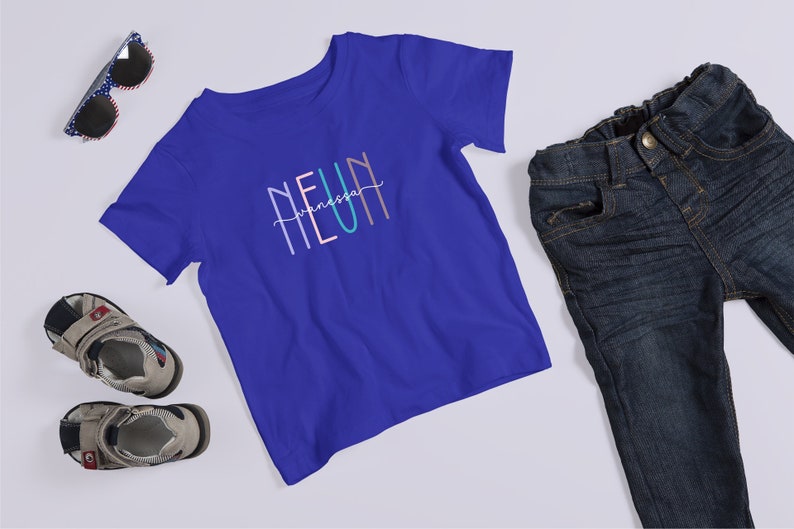 May include: A royal blue short-sleeve t-shirt featuring the text NEUN with the name Vanessa written in cursive script across the center. The shirt is displayed alongside a pair of dark wash denim jeans, gray and black sandals, and sunglasses.