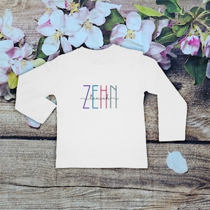 May include: A white long-sleeve shirt featuring the word ZEHIN in vertical, multi-colored block letters with the word taniah written in a black cursive script across the center of the block letters, creating a layered graphic design.