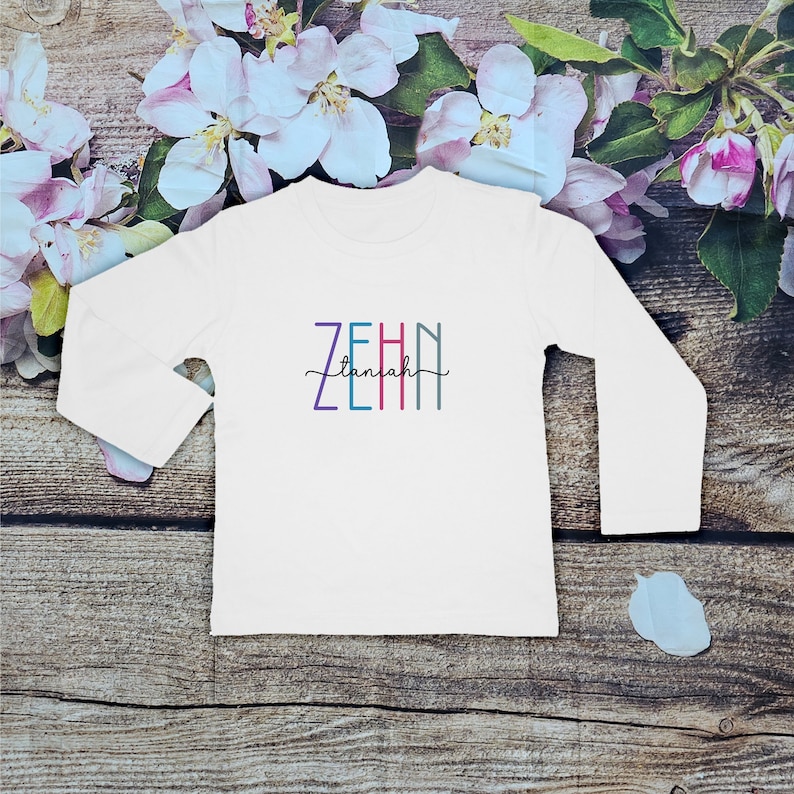 May include: A white long-sleeve shirt featuring the word ZEHIN in vertical, multi-colored block letters with the word taniah written in a black cursive script across the center of the block letters, creating a layered graphic design.