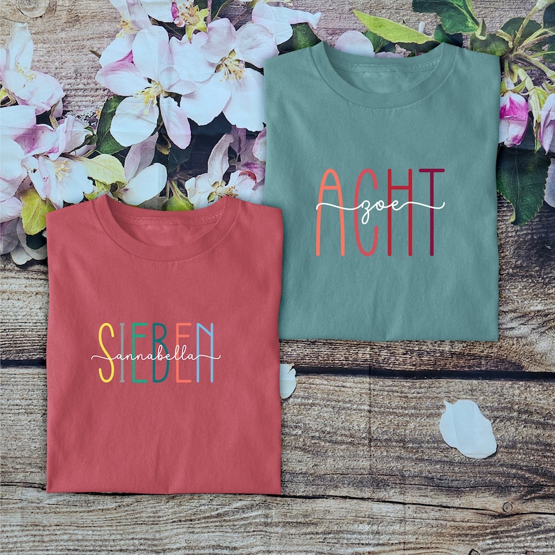 May include: A red t-shirt with the text "SIEBEN" and "annabella" in colorful letters, and a teal t-shirt with the text "ACHT" and "zoe" in colorful letters. Both shirts feature a crew neckline and short sleeves, displayed as folded garments.