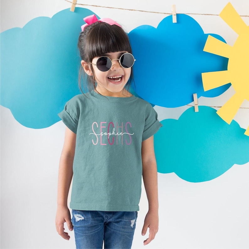 Sophie trägt unser Geburtstagsshirt für Mädchen und Jungen in Atlantic mit einer Sechs in Blassrosa - Rosa - Pink - Altrosa - Berry