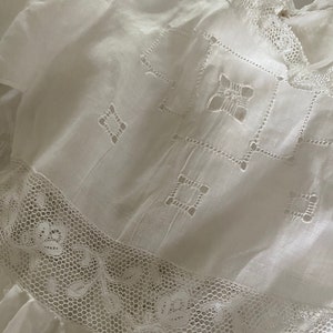 Op de afbeelding: Witte katoenen jurk met delicate kantversiering en geborduurde details. De jurk heeft een vierkant patroon met kleine gaatjes in de hele stof.