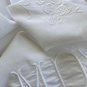 以下が含まれることがあります： 白いリネン寝具、刺繍の花柄と文字のデザイン。 刺繍は、繊細な蝶と葉の模様と、エレガントなスクリプト文字が特徴です。 折りたたまれたリネンは、複雑なディテールを示す、重ねられた効果を生み出しています。