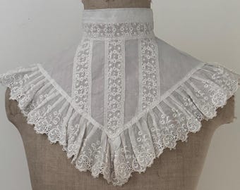May include: A white, vintage-style collar with intricate lace and floral embroidery. The collar features a high, rounded neckline and a ruffled, scalloped edge. The design includes vertical panels of detailed stitching.