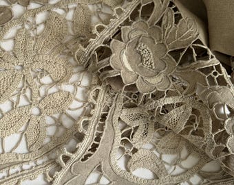 May include: A beige crocheted lace tablecloth with a floral design. The lace is delicate and intricate, with a scalloped edge.