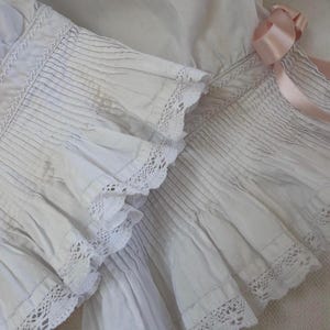 May include: A white garment featuring intricate smocking details and delicate white lace trim along the ruffled edges. A soft, pale pink satin ribbon is tied in a bow on the side, adding a subtle contrast to the textured, pleated fabric construction.