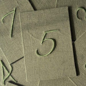 May include: A collection of rectangular olive green fabric cards, each featuring a single embroidered number in a lighter shade of green. The central card prominently displays the number five, while other cards show partial numbers.