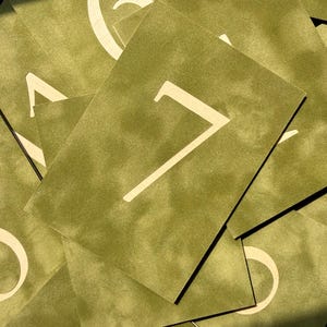 May include: A collection of rectangular olive green cards featuring large, light-colored numerals printed in the center. The cards are layered on top of each other, displaying various numbers including a prominent seven in the foreground.