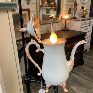 Può includere: Una lampada a forma di teiera in stile vintage, di colore azzurro chiaro con dettagli dorati. La lampada presenta un beccuccio, un manico e piedini decorativi, con una lampadina calda e luminosa all'interno. La lampada è montata su un supporto in metallo nero.