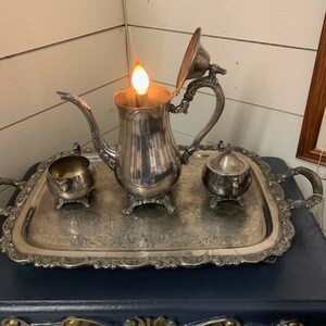 May include: A silver tea set with a lit candle inside the teapot, placed on a decorative tray. The set includes a teapot, sugar bowl, and creamer. The tray has ornate edges and handles, resting on a dark blue table.