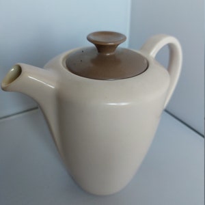 May include: A cream-colored ceramic teapot with a brown lid and a brown knob on top. The teapot has a spout and a handle.