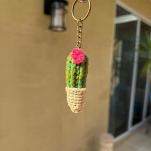 May include: A handmade cactus keychain. The cactus is green and yellow with a pink flower on top, and sits in a beige pot. The keychain is attached to a gold-colored ring and chain.