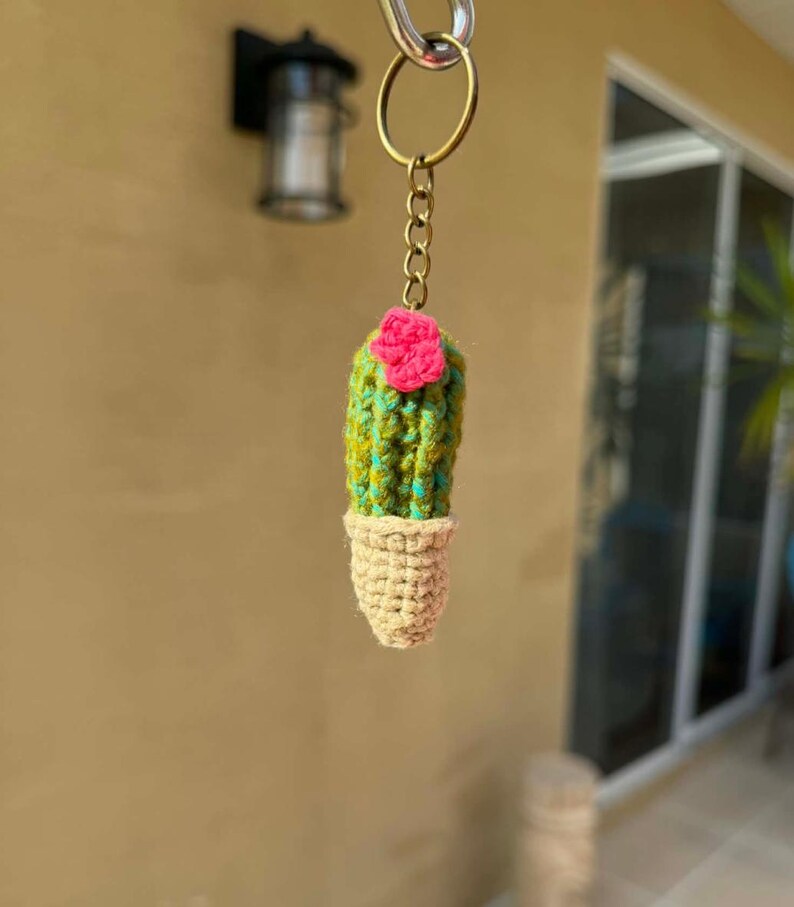 May include: A handmade cactus keychain. The cactus is green and yellow with a pink flower on top, and sits in a beige pot. The keychain is attached to a gold-colored ring and chain.