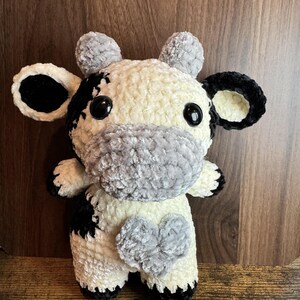 Peut inclure: Une peluche faite &agrave; la main en forme de vache, &agrave; l'aspect doux et textur&eacute;. La vache pr&eacute;sente un motif noir et blanc, avec des d&eacute;tails noirs sur les oreilles, les sabots et les taches. Elle a un museau gris et une tache en forme de c&oelig;ur gris sur la poitrine. Les yeux sont noirs.