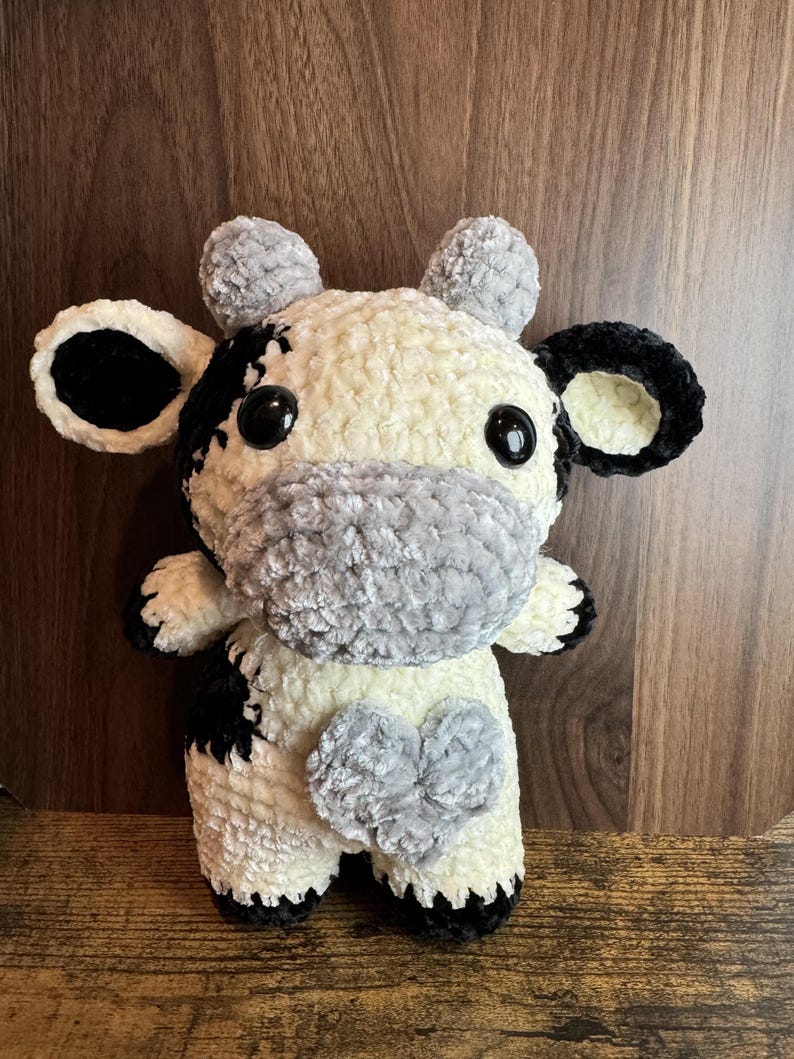 Peut inclure: Une peluche faite &agrave; la main en forme de vache, &agrave; l'aspect doux et textur&eacute;. La vache pr&eacute;sente un motif noir et blanc, avec des d&eacute;tails noirs sur les oreilles, les sabots et les taches. Elle a un museau gris et une tache en forme de c&oelig;ur gris sur la poitrine. Les yeux sont noirs.