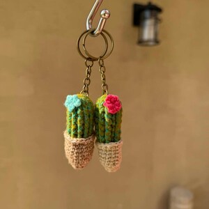 May include: Two handmade crochet cactus keychains. Each cactus is green and yellow with a small flower on top, one blue and one pink. The cacti are in tan-colored pots. The keychains hang from a metal ring.