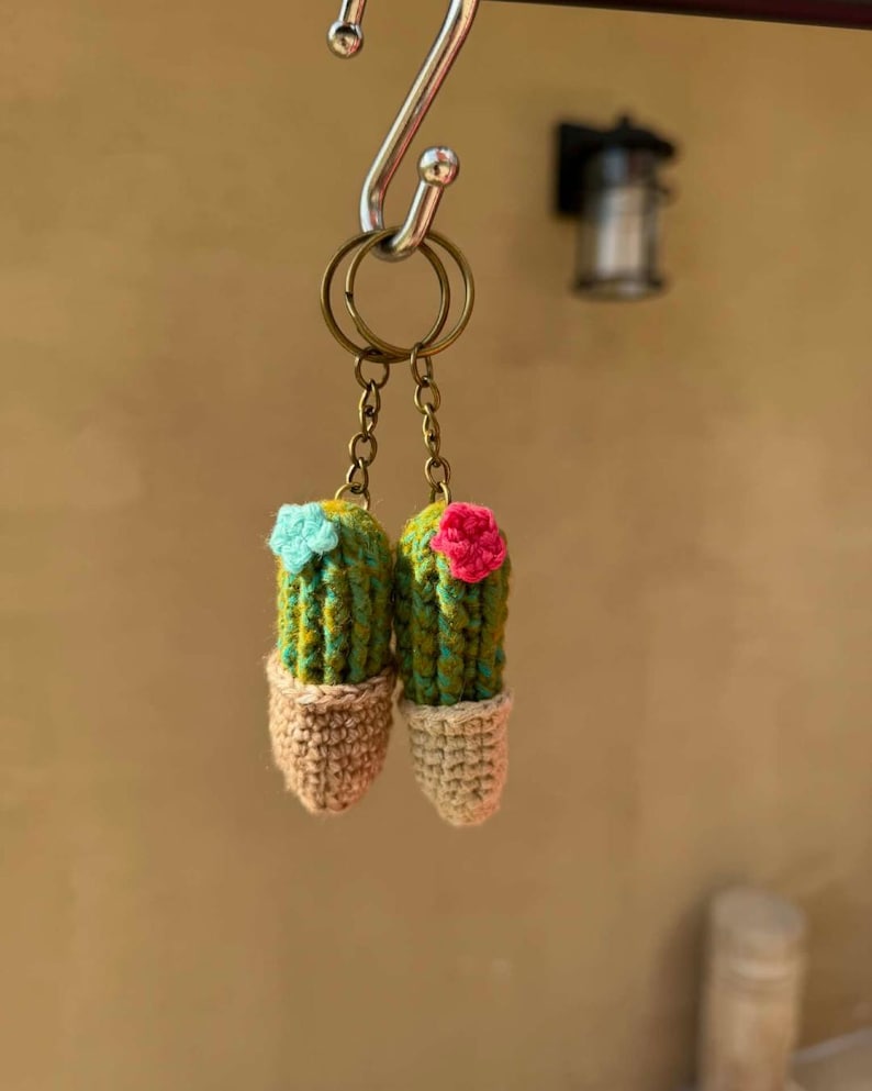 May include: Two handmade crochet cactus keychains. Each cactus is green and yellow with a small flower on top, one blue and one pink. The cacti are in tan-colored pots. The keychains hang from a metal ring.