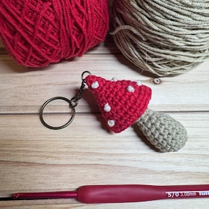 Peut inclure: Un porte-cl&eacute;s champignon crochet&eacute; &agrave; la main, avec un chapeau rouge et des pois blancs. Deux pelotes de laine, une rouge et une beige, sont en arri&egrave;re-plan, ainsi qu'un crochet de 3,00 mm.
