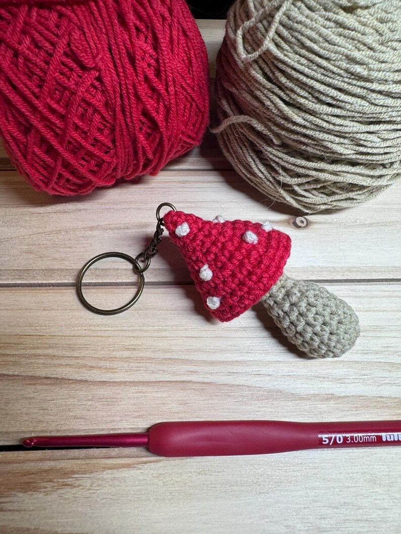 Peut inclure: Un porte-cl&eacute;s champignon crochet&eacute; &agrave; la main, avec un chapeau rouge et des pois blancs. Deux pelotes de laine, une rouge et une beige, sont en arri&egrave;re-plan, ainsi qu'un crochet de 3,00 mm.