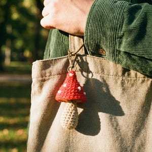 Peut inclure: Un porte-cl&eacute;s en forme de champignon fait &agrave; la main, avec un chapeau rouge &agrave; pois blancs et un pied beige. Le champignon est accroch&eacute; &agrave; un fermoir en m&eacute;tal et suspendu &agrave; un sac fourre-tout beige. En arri&egrave;re-plan, une personne porte une veste en velours c&ocirc;tel&eacute; verte.