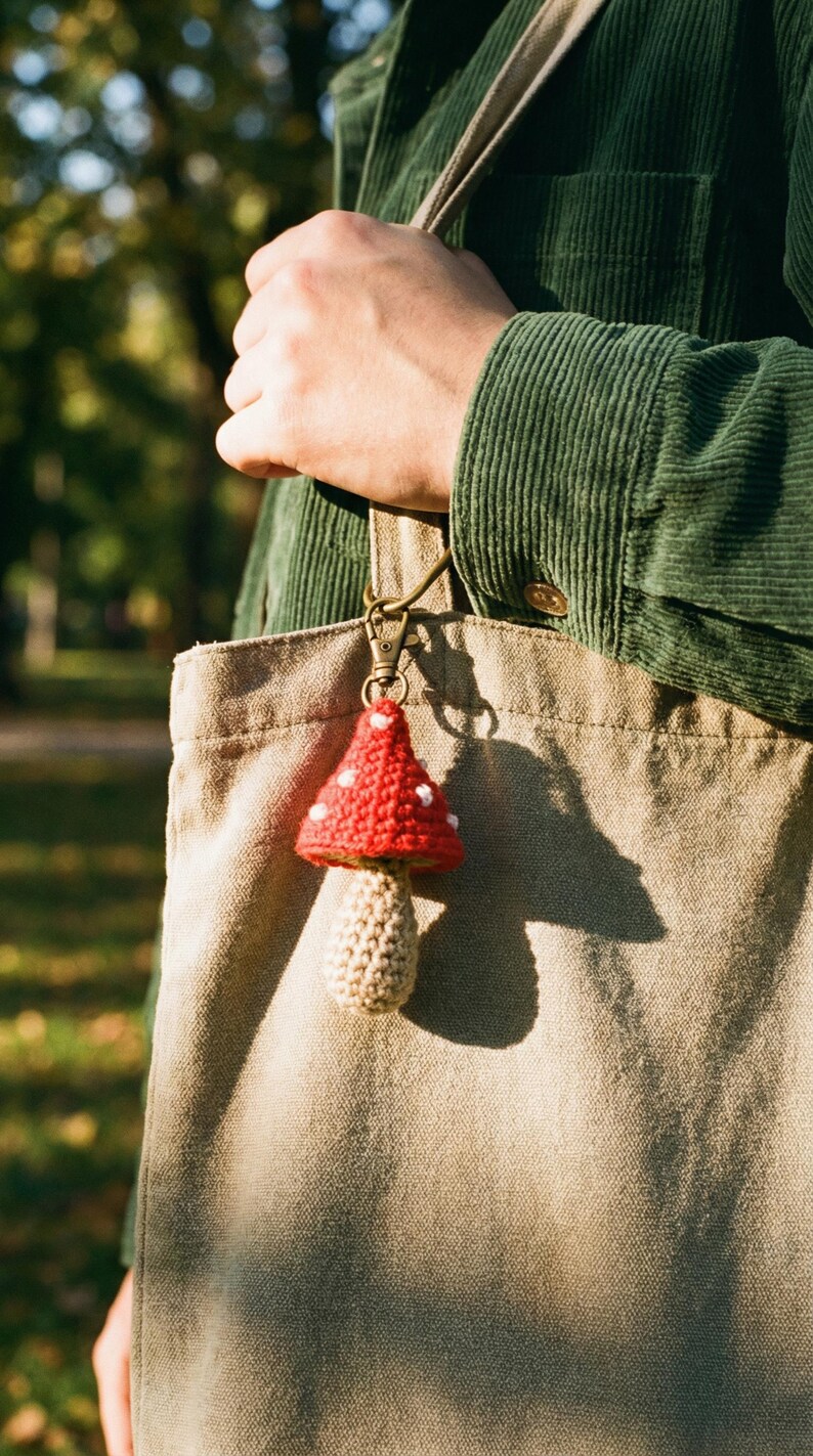 Peut inclure: Un porte-cl&eacute;s en forme de champignon fait &agrave; la main, avec un chapeau rouge &agrave; pois blancs et un pied beige. Le champignon est accroch&eacute; &agrave; un fermoir en m&eacute;tal et suspendu &agrave; un sac fourre-tout beige. En arri&egrave;re-plan, une personne porte une veste en velours c&ocirc;tel&eacute; verte.