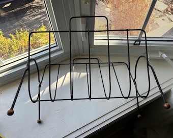 May include: Black metal magazine rack with rounded edges and four legs with ball-shaped ends. The rack is sitting on a white windowsill.