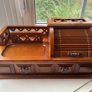 May include: A vintage wooden desk organizer with a warm brown finish. It features a roll-top compartment, two small drawers with metal pulls, and an open tray area. The design includes decorative cutouts and a small railing.