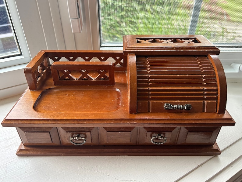 May include: A vintage wooden desk organizer with a warm brown finish. It features a roll-top compartment, two small drawers with metal pulls, and an open tray area. The design includes decorative cutouts and a small railing.