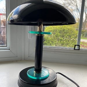 Könnte beinhalten: Eine schwarze Schreibtischlampe mit einem kuppelförmigen Schirm und einem grünen Glasfuß. Die Lampe hat einen goldfarbenen Akzent auf der Oberseite des Schirms und um den Fuß herum.