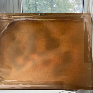 May include: The back of a framed picture with a brown paper backing and a clear plastic covering. The frame is made of wood and has a light brown finish.